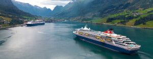 Two cruise ships navigating a scenic fjord surrounded by green mountains and snow-capped peaks, representing ALMACO’s turnkey marine outfitting and accommodation solutions for cruise and expedition vessels.