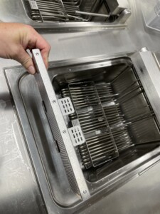 Hand holding a metal filter above a stainless steel container with parallel metal rods, part of an industrial kitchen appliance designed by ALMACO for marine and offshore food service systems.