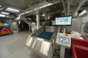 Industrial laundry facility with conveyor system, colorful laundry bags, and control monitors – part of ALMACO’s turnkey laundry and automation solutions for cruise ships, offshore platforms, and marine accommodation.