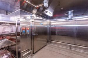 Interior of a stainless steel walk-in refrigerator with control panels, thermometer, and food storage shelves, designed for marine galley cold storage and offshore catering environments.