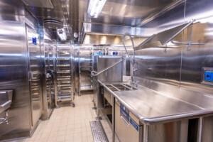 Stainless steel commercial galley kitchen with industrial sinks, mobile storage racks, and hygienic tiled flooring, designed for marine and offshore food service operations.