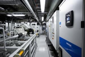 ALMACO mechanical room featuring Vacon control panels, overhead piping, and industrial systems. Engineered for marine and offshore applications with safety railings and efficient layout for shipboard environments.