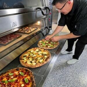 Person preparing pizzas in front of industrial oven with metal rack and tiled floor – ALMACO marine galley and foodservice equipment