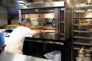 Person placing pizza into PizzaMaster commercial oven with digital temperature display and trays of ingredients – ALMACO marine galley and foodservice equipment