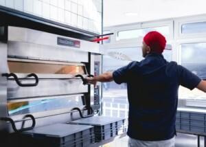 Chef operating PizzaMaster commercial oven in professional galley kitchen with stacked trays – marine catering equipment by ALMACO