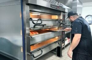 Chef operating a PizzaMaster oven in a professional marine galley, baking pizza in a stainless steel commercial kitchen – ALMACO marine catering solutions and food service equipment
