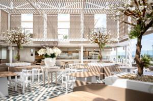 Outdoor dining area with wooden tables, white chairs, and potted flowering trees under slatted ceiling – ALMACO marine hospitality and modular foodservice design