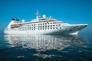 White cruise ship with multiple decks and lifeboats sailing on calm reflective waters under a clear blue sky, showcasing ALMACO’s turnkey marine accommodation and modular outfitting solutions.